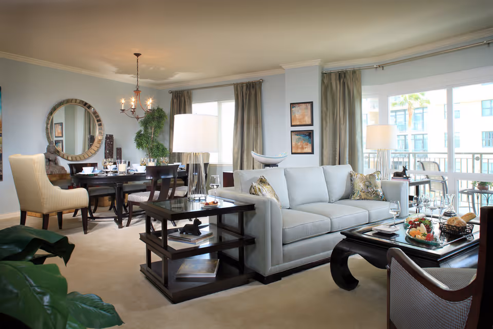 A bright and spacious living room and dining area in a senior living facility. The living room features a light gray sofa with patterned throw pillows, a dark wooden coffee table with a tray of snacks and wine glasses, and a side table with a lamp. The dining area has a round dark wooden table set with dishes and glasses, surrounded by chairs including a beige upholstered armchair. Large windows with curtains allow natural light to fill the room, and a decorative round mirror hangs on the wall above a small console table with a statue and plants.