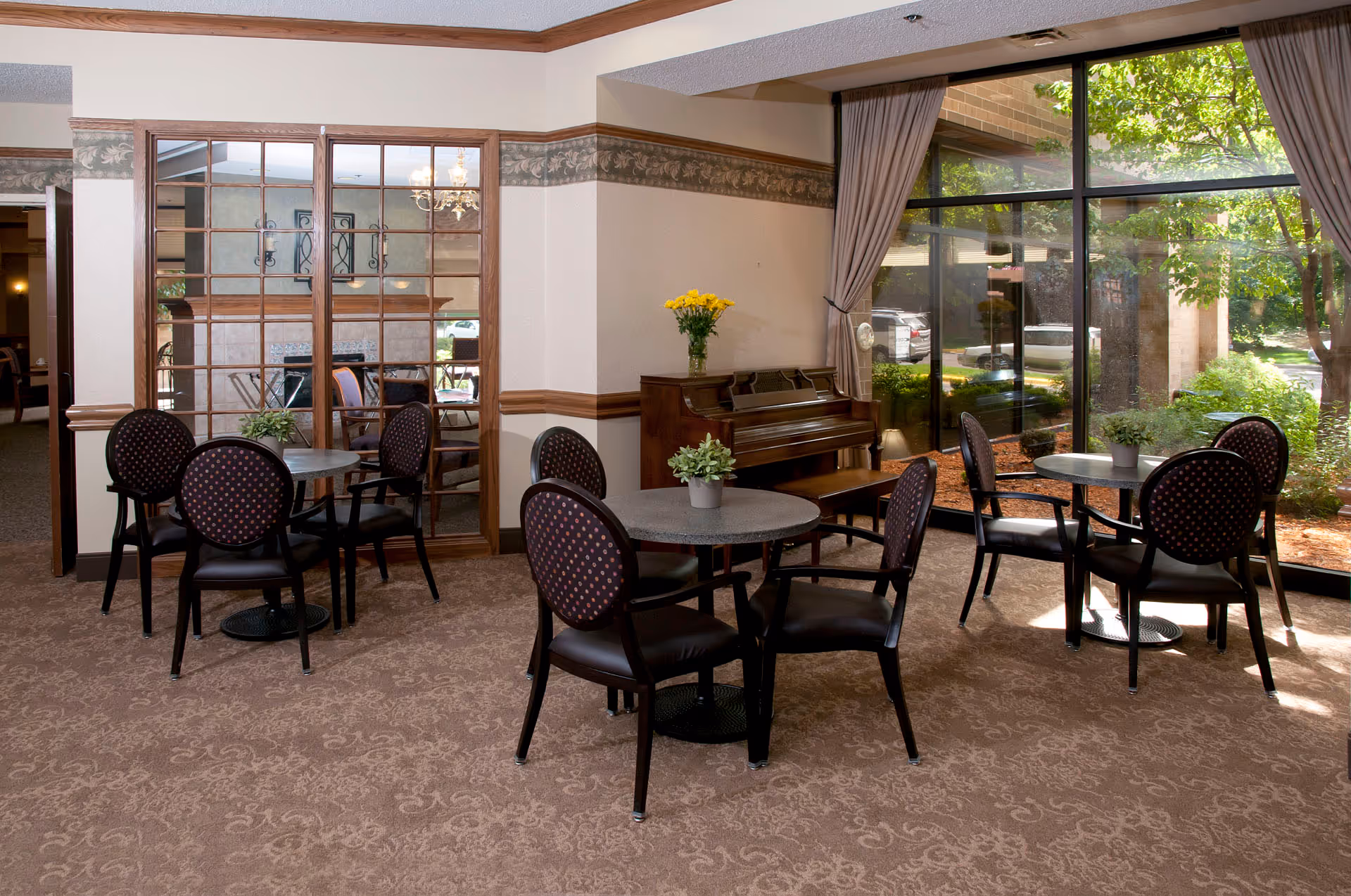 Sunlit common room with round tables and chairs, a piano, and large windows overlooking a garden.