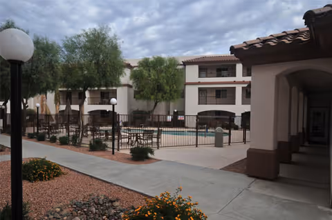 Outdoor courtyard area of a retirement home featuring a fenced swimming pool surrounded by patio tables with umbrellas, trees, and a three-story building with balconies in the background under a cloudy sky.