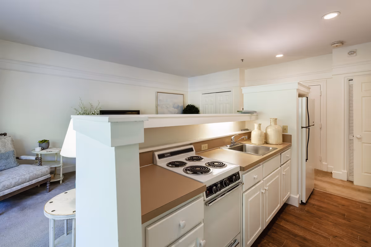 Interior view of a small kitchen area in a retirement residence featuring a stove with four coil burners, a sink with two decorative vases on the countertop, white cabinetry, and a refrigerator. Adjacent to the kitchen is a living area with a cushioned chair, a small round table, and a lamp. The walls are painted white, and the floor transitions from wood in the kitchen to carpet in the living area.