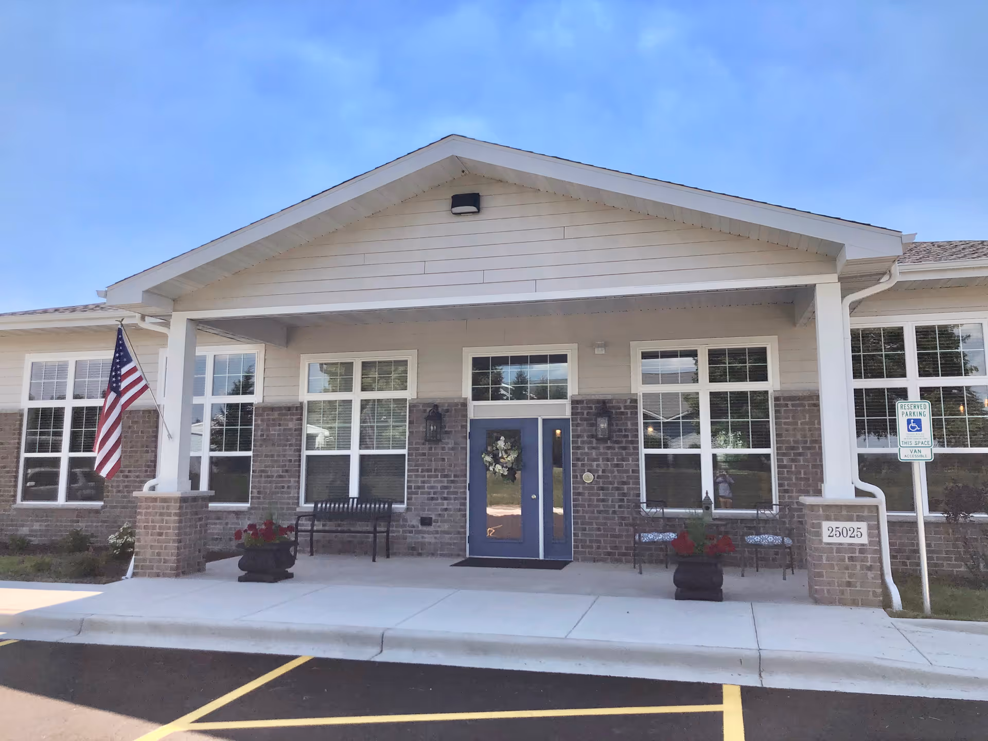 Front exterior view of a single-story senior living facility building with a covered entrance, brick and siding facade, large windows, an American flag, benches, potted plants, and a reserved parking sign for handicapped access.