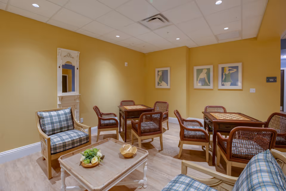 A cozy common area with yellow walls featuring two small wooden tables with built-in chessboards, surrounded by six wooden chairs with woven backs and seats. In the foreground, there are two cushioned armchairs with plaid upholstery and a wooden coffee table with decorative items. Three framed artworks of stylized figures hang on the wall.