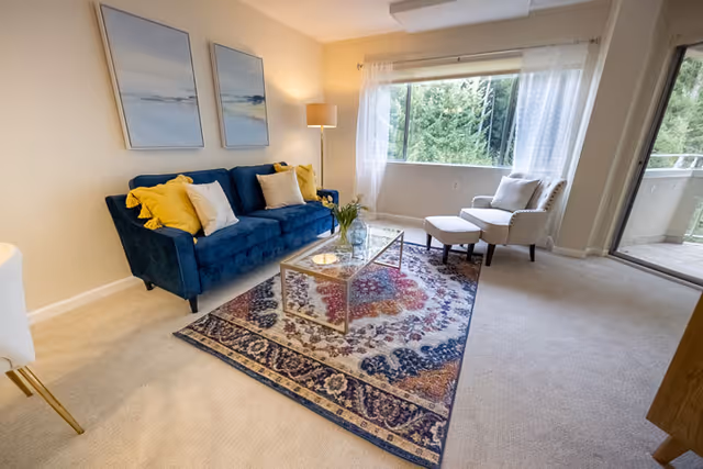 A bright living room with a large window showing green trees outside. The room features a blue sofa with yellow and white pillows, a glass coffee table on a patterned rug, a white armchair with an ottoman, a floor lamp, and light-colored carpet. Sheer white curtains hang on the window, and a sliding glass door leads to a balcony.