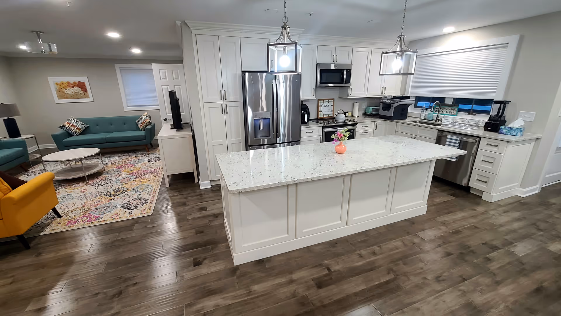 A modern open-concept kitchen and living room area with white cabinetry, stainless steel appliances, and a large white island countertop with a small flower vase. The living room has teal sofas, a yellow armchair, a round coffee table, and a colorful patterned rug on wooden flooring.