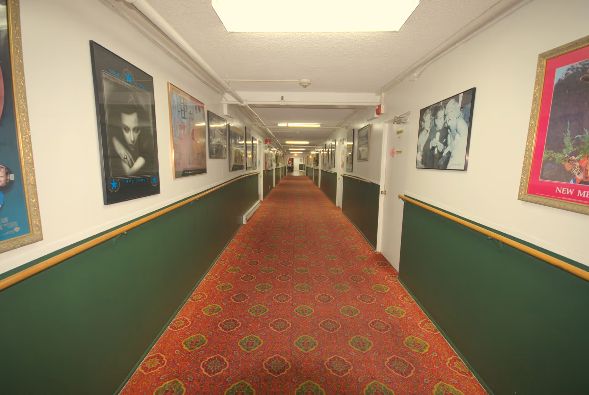 Long carpeted corridor in a senior living facility lined with framed pictures and handrails on both sides.