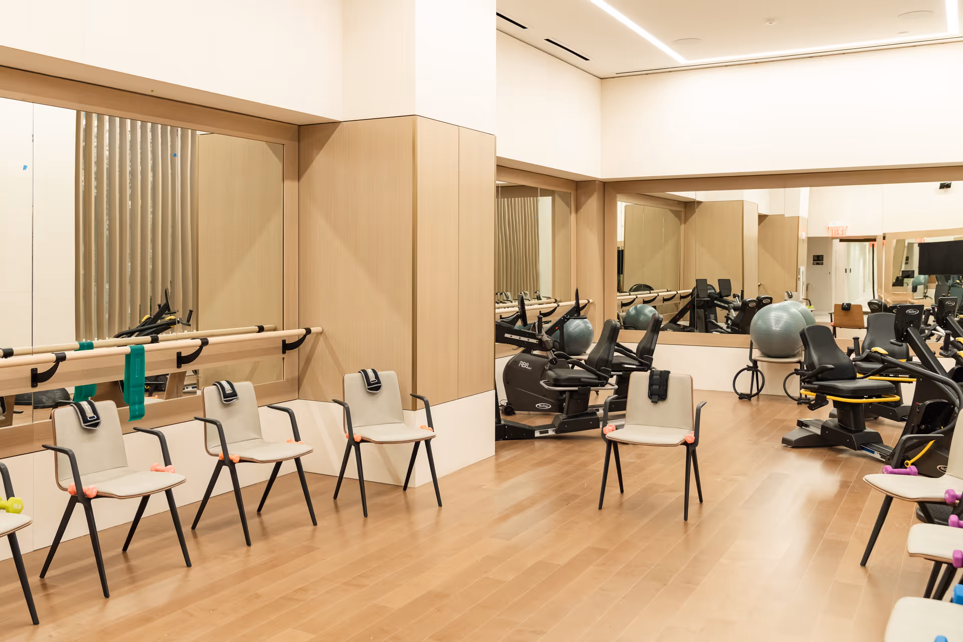 A fitness room with wooden floors and light-colored walls featuring large mirrors. Several chairs are arranged in a semi-circle, each with small exercise weights on the seats. The room also contains exercise equipment including stationary bikes and large exercise balls.