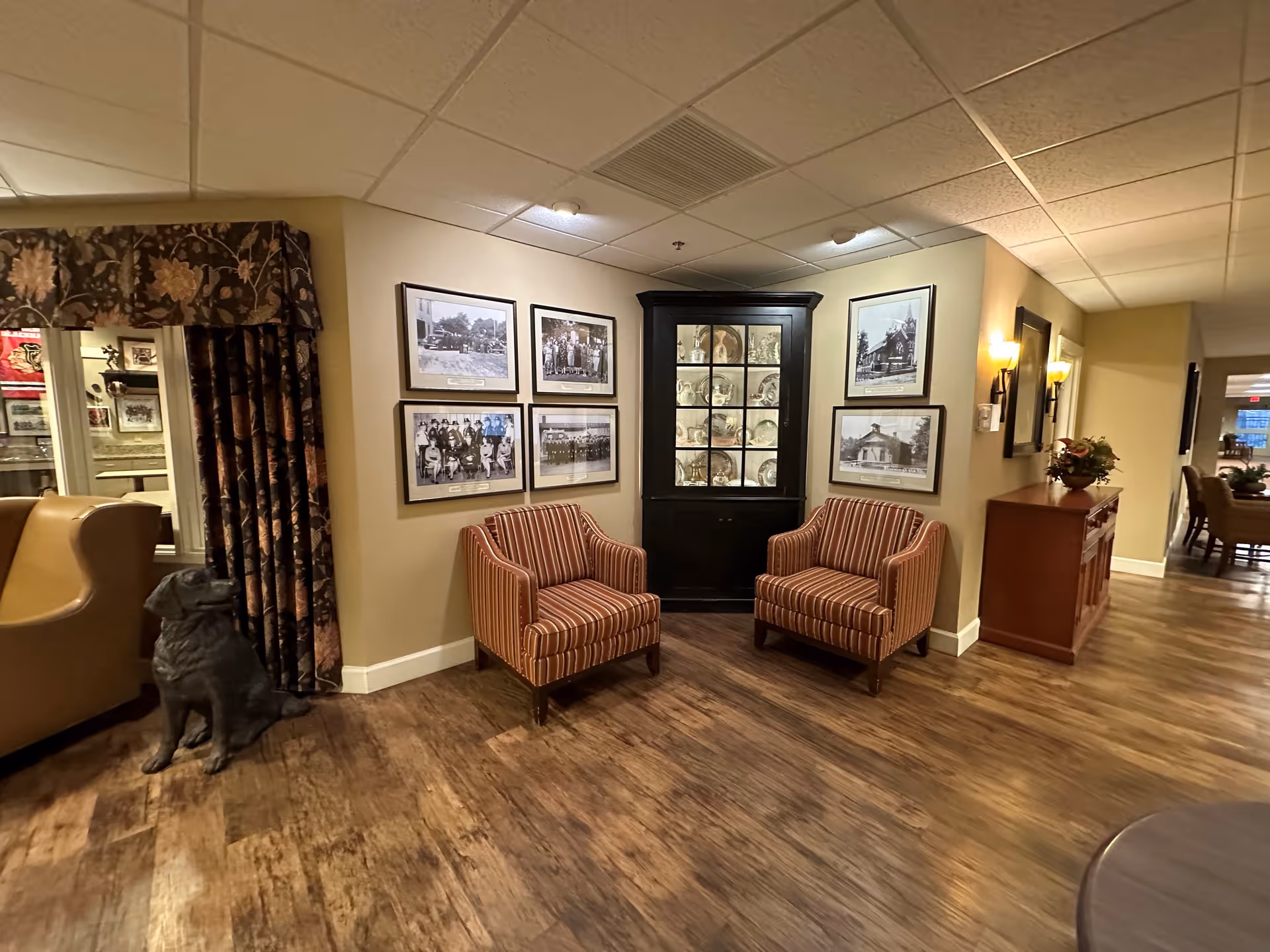 A cozy seating area in a senior living facility with two striped armchairs placed in front of a black corner cabinet displaying decorative plates. The walls are adorned with framed black and white photographs. To the left, there is a window with floral curtains and a statue of a dog on the floor. The floor is wooden, and the lighting is warm with wall sconces and ceiling lights.