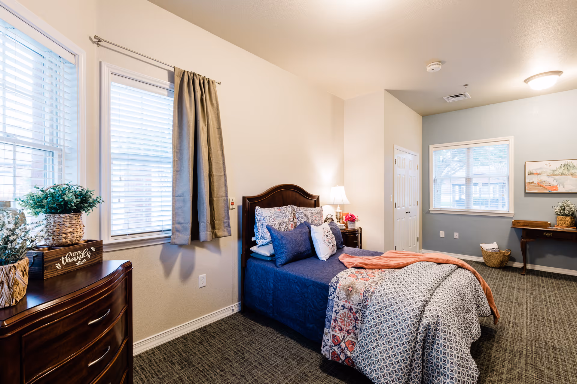 A well-lit bedroom with a neatly made bed featuring blue and patterned bedding. There is a wooden dresser with plants and a decorative 'Give Thanks' sign on it. The room has two windows with blinds and curtains, a nightstand with a lamp and flowers, a small desk with a basket underneath, and a framed picture on the wall.