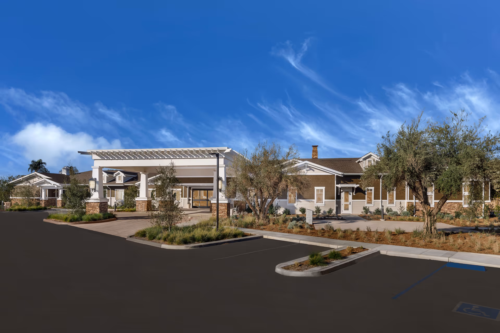 Exterior view of Clearwater at North Tustin senior living facility showing a single-story building with a covered entrance, landscaped grounds with trees and shrubs, and a parking lot with marked spaces including a handicapped spot under a clear blue sky.