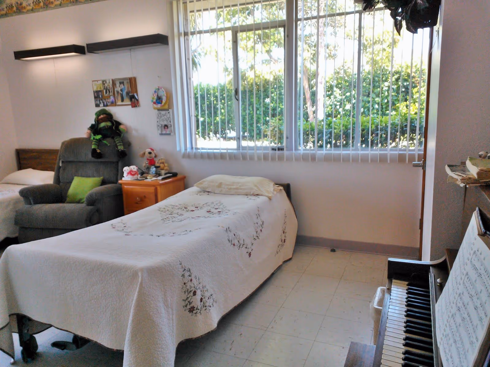 A bright bedroom with a single bed covered in a white quilt with floral patterns. Next to the bed is a wooden nightstand with stuffed animals and a phone on it. There is a gray recliner chair with a green pillow and a doll sitting on it. A large window with vertical blinds lets in natural light and shows greenery outside. A piano with sheet music is visible on the right side of the room.