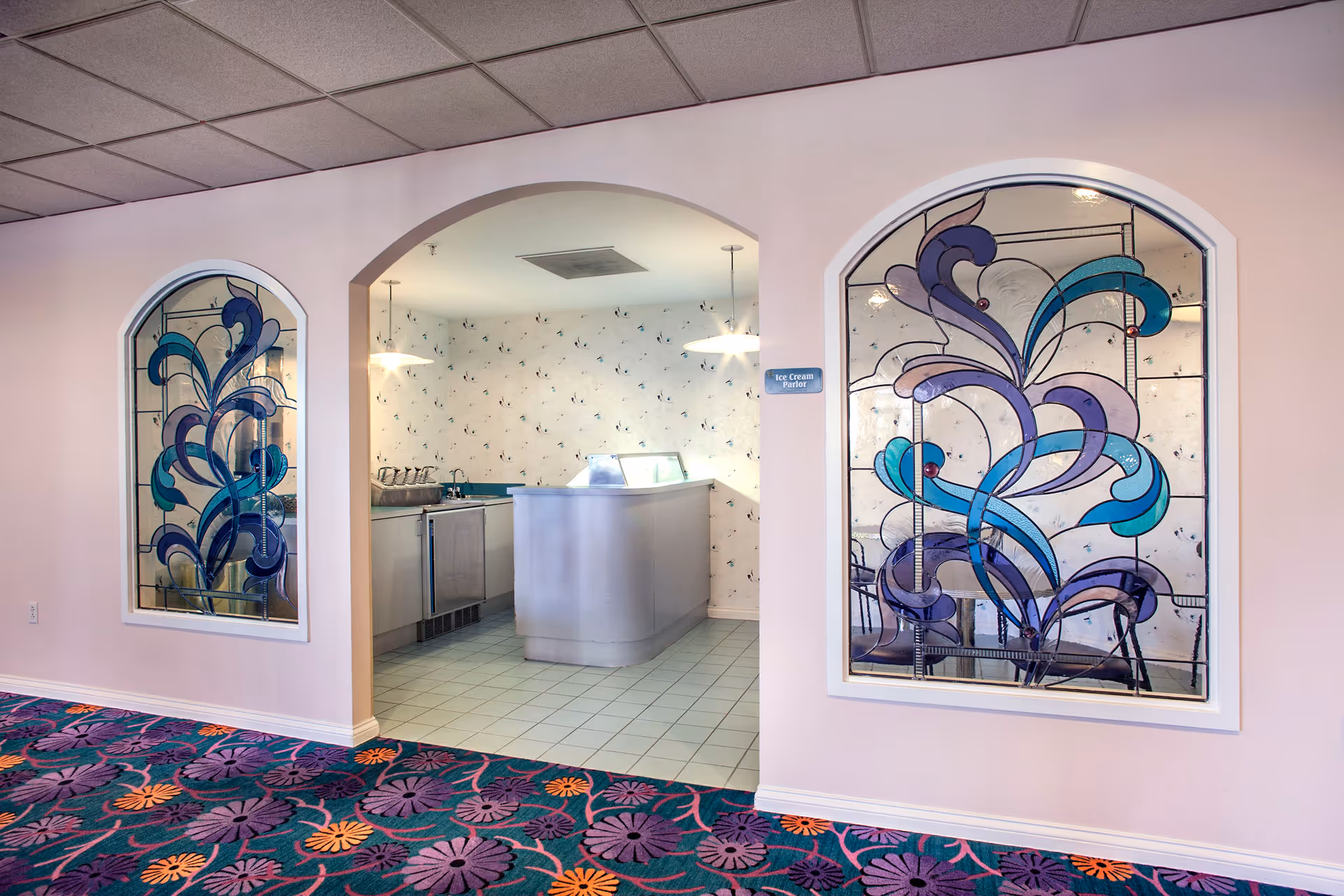 Interior view of an ice cream parlor area in a senior living facility. The room features an arched entrance with two decorative stained glass windows on either side, showcasing blue and purple abstract floral designs. Inside, there is a silver ice cream counter and a tiled floor. The walls have a light wallpaper with a subtle pattern, and two pendant lights hang from the ceiling. The carpet outside the parlor has a colorful floral pattern in purple, orange, and green tones.