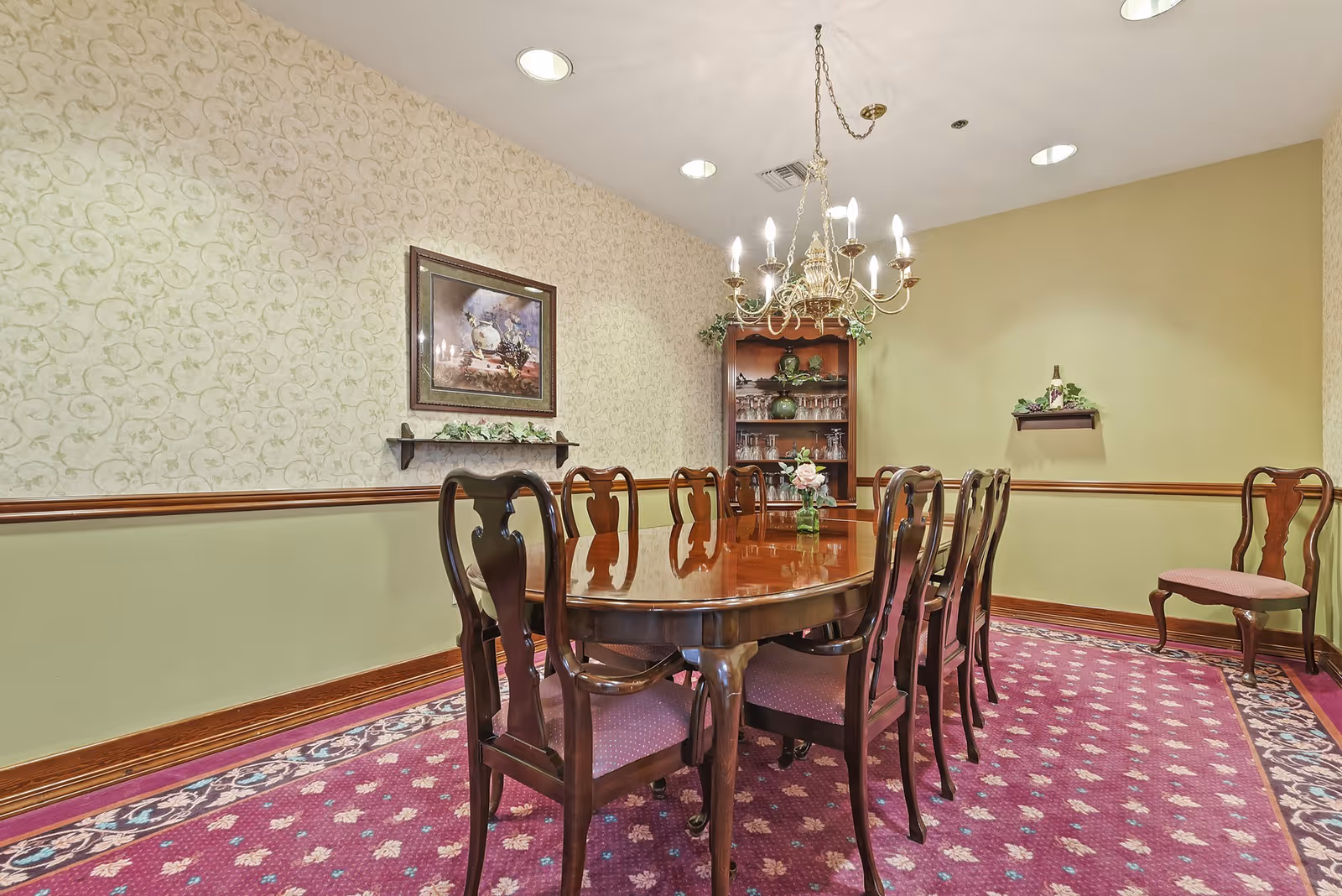 A formal dining room with a polished wooden dining table surrounded by eight matching wooden chairs with cushioned seats. The room features a patterned purple carpet, light green walls with a decorative chair rail, a chandelier hanging above the table, a framed painting on the wall, and a wooden cabinet with glassware in the corner.