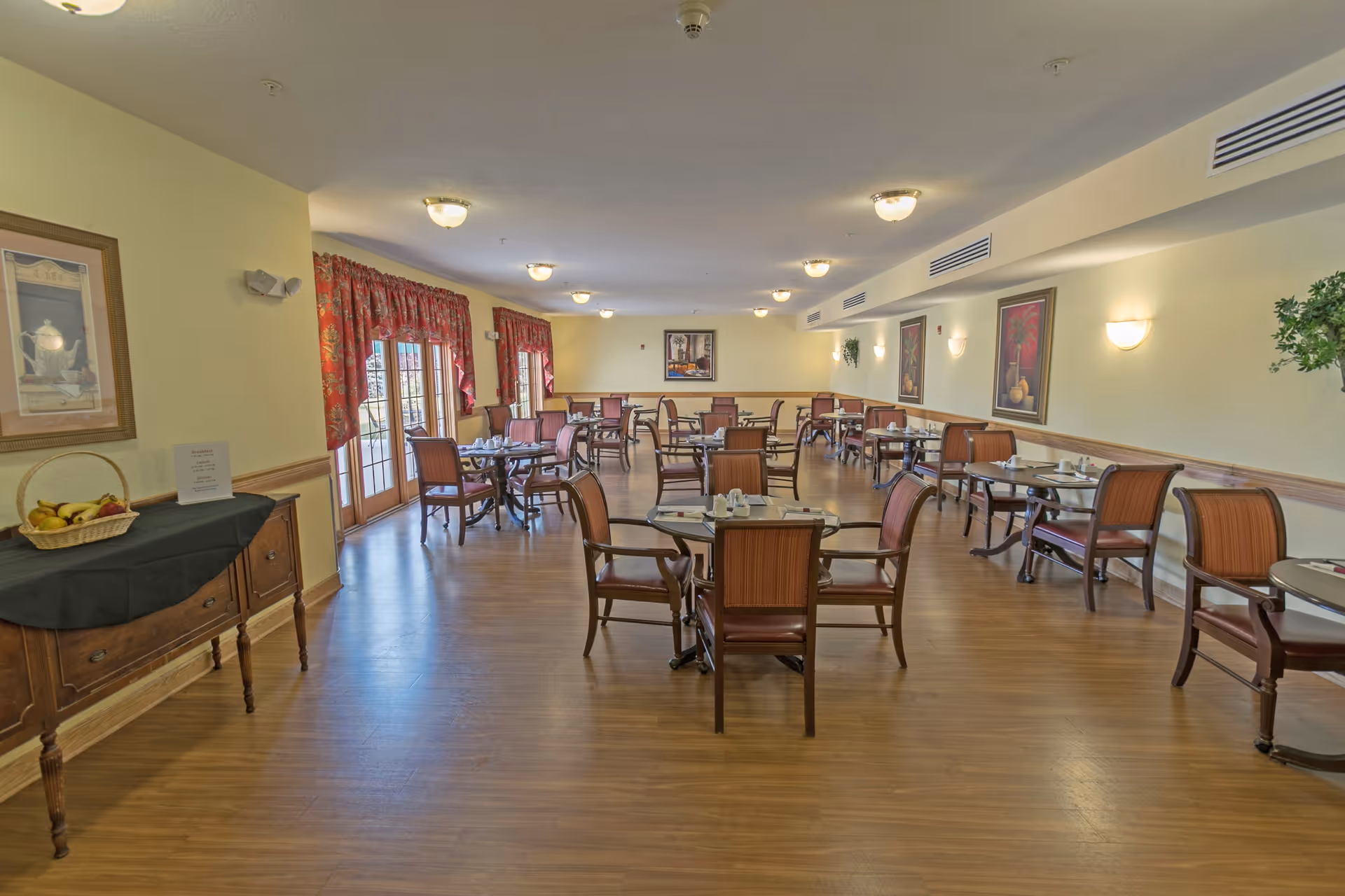 A spacious dining room with multiple wooden tables and chairs arranged neatly. The room has wooden flooring, light yellow walls, framed artwork, and red patterned curtains on the windows. A side table with a basket of fruit and a sign is visible on the left side.