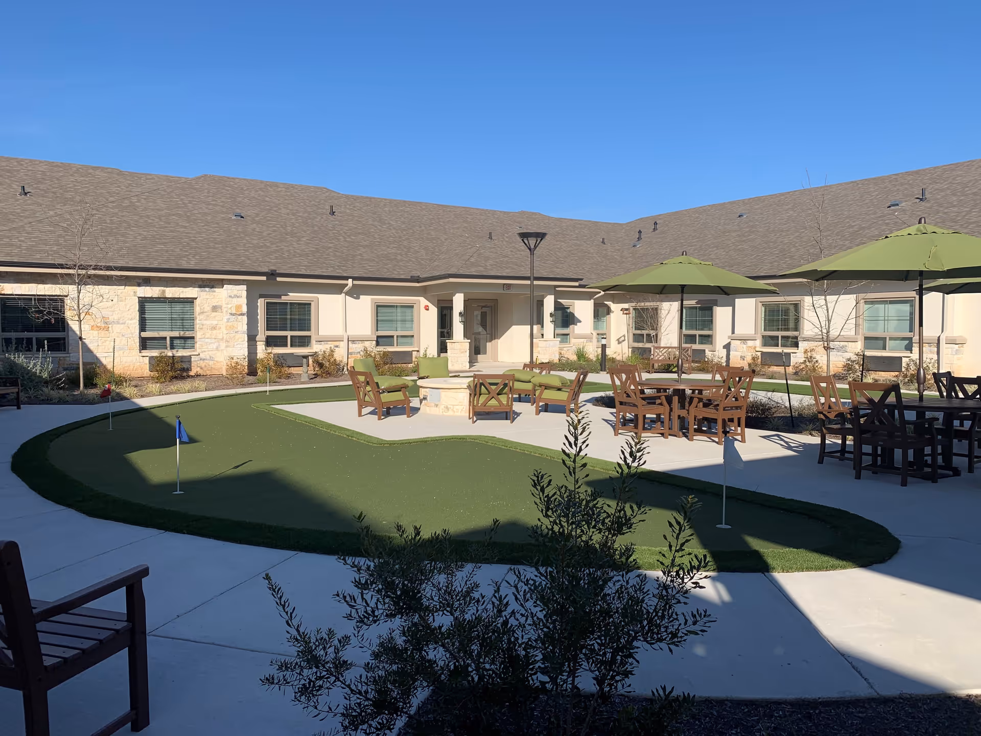 Outdoor courtyard area at Volante Senior Living of Sage Spring featuring a putting green with small flags, surrounded by concrete walkways. There are several wooden chairs and tables with green umbrellas, and a fire pit with cushioned seating in the center. The building with beige walls and multiple windows encloses the courtyard under a clear blue sky.