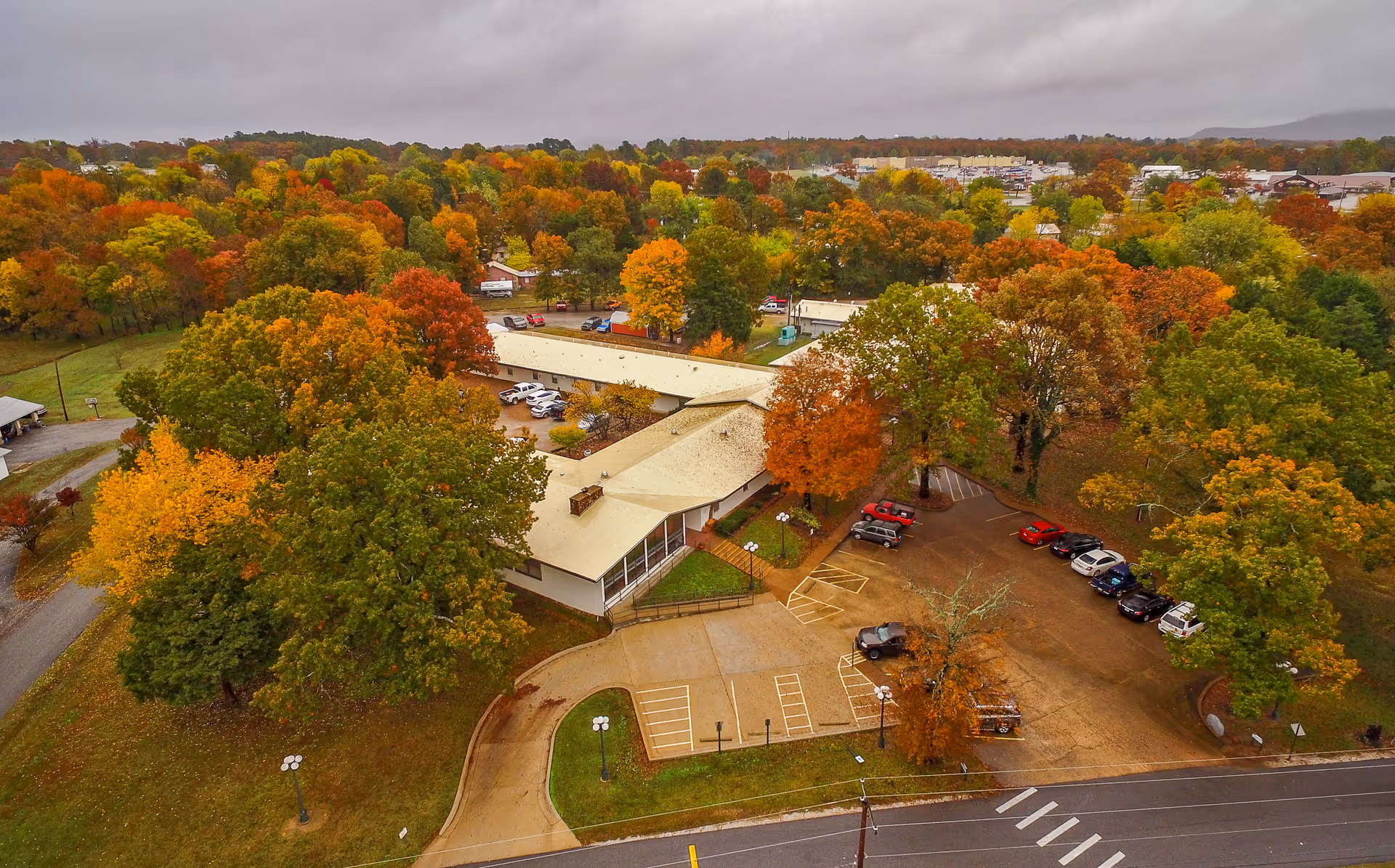 Aerial view of The Blossoms at Mountain View Rehab & Nursing Center surrounded by trees with autumn foliage. The building has a light-colored roof and is situated near a parking lot with several cars. The surrounding area includes a mix of green, yellow, orange, and red trees under a cloudy sky.