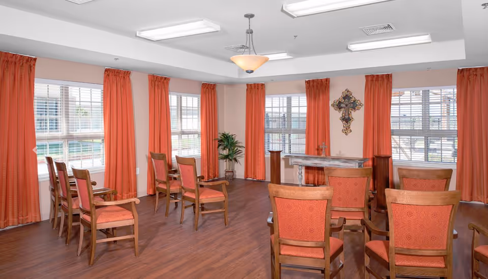 A room with wooden chairs arranged in rows facing a small altar table with a decorative cross mounted on the wall behind it. The room has large windows with white blinds and orange curtains, wood flooring, and a ceiling light fixture.