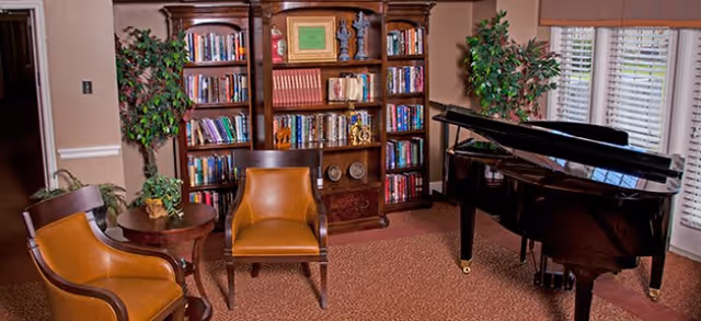 Cozy common room with a grand piano, a large bookshelf, two leather chairs, a side table, and potted plants.