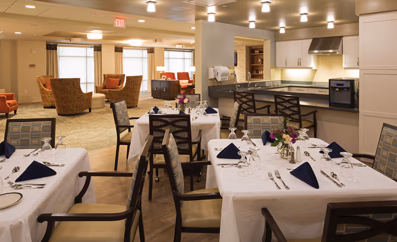 A dining area in a senior living facility with tables covered in white tablecloths, set with silverware, glasses, and navy blue folded napkins. There are floral centerpieces on the tables. In the background, there is a kitchen counter with bar stools and a seating area with armchairs and a sofa near large windows.