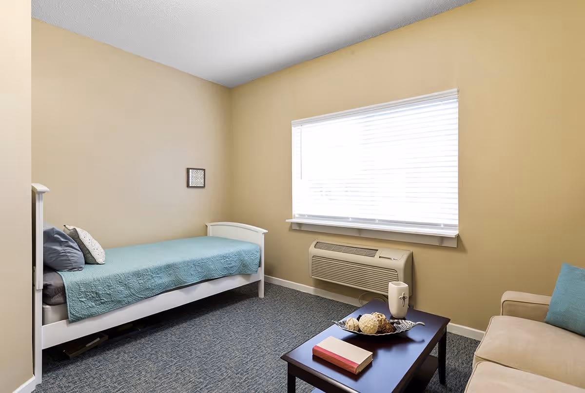 A small, simple bedroom with beige walls and a window with white blinds. The room contains a single white bed with a teal quilt and pillows, a beige sofa with a teal cushion, and a dark wooden coffee table with decorative items and a book. Below the window is a wall-mounted air conditioning unit.