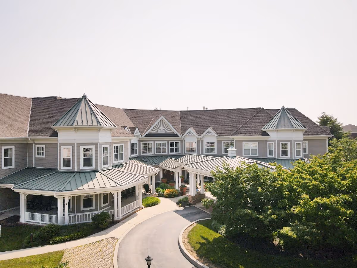 Exterior view of a senior living facility building with a circular driveway, two prominent turret-like structures with green metal roofs, multiple windows, and surrounding greenery including trees and shrubs.