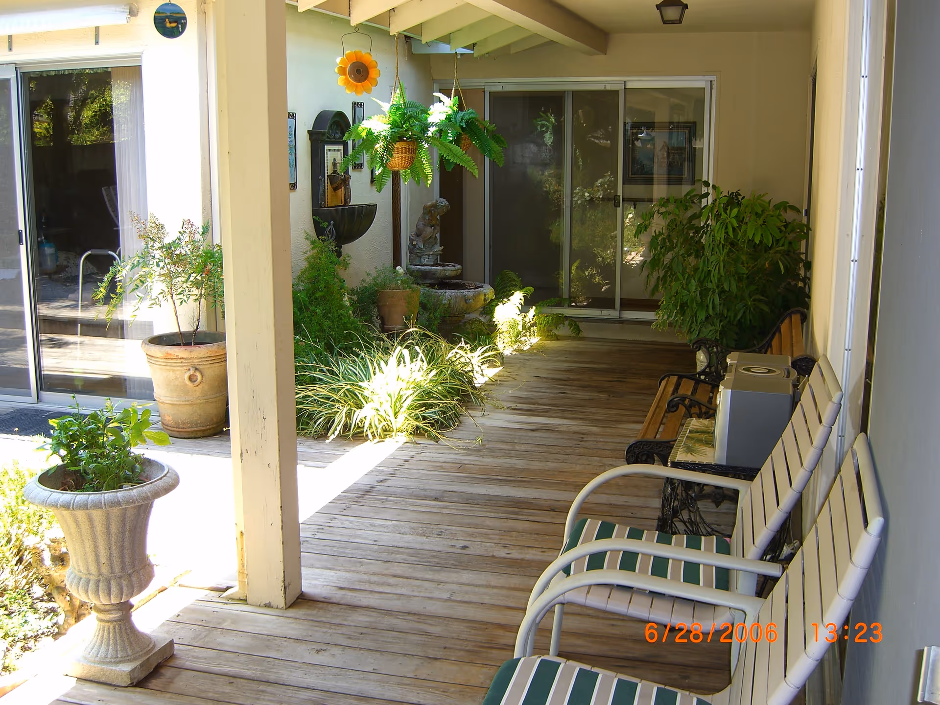 Covered wooden porch area with several potted plants, hanging fern, a small water fountain, and two white chairs with green and white striped cushions. Sliding glass doors lead into the building, and there is a bench with a small box on it along the wall.