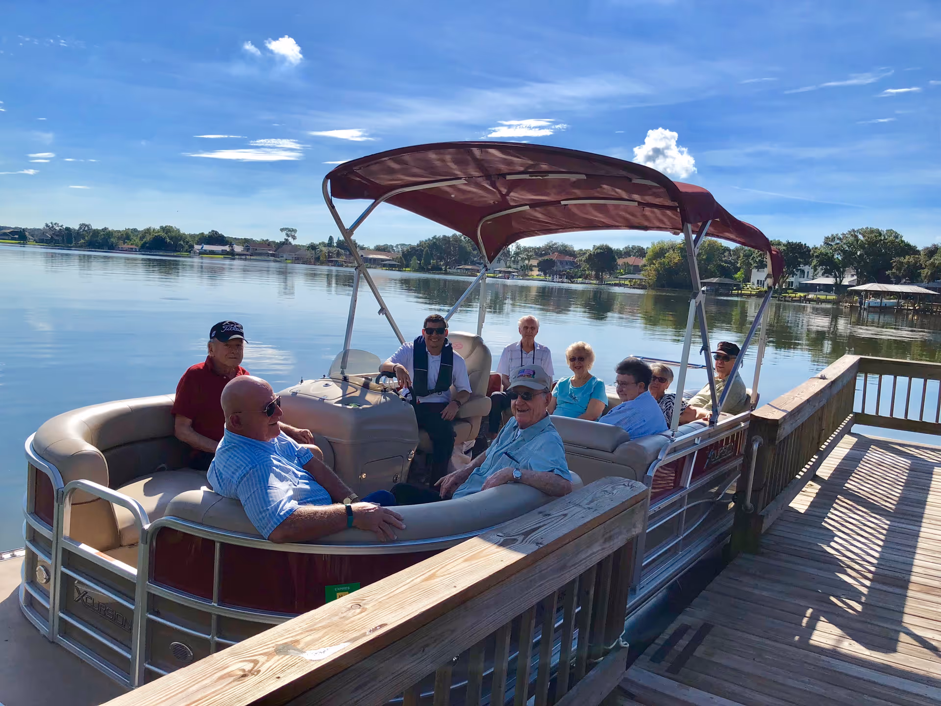 A group of elderly people sitting and enjoying a boat ride on a calm lake under a clear blue sky, with houses and trees visible in the background.