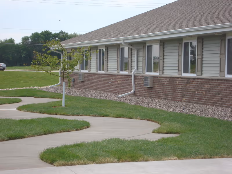 Exterior view of a single-story building with beige siding and brick lower walls, several windows, a small tree, and a curved concrete walkway surrounded by green grass.