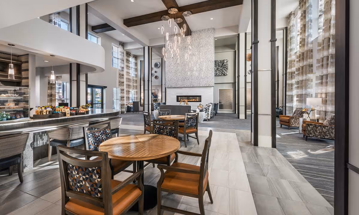 A spacious and modern common area in a senior living facility featuring round wooden tables with chairs, a long counter with stools, a contemporary fireplace, high ceilings with exposed beams, large windows with sheer curtains, and comfortable seating areas with armchairs.