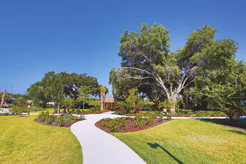 A bright outdoor garden area with a winding concrete pathway surrounded by green grass, various trees, and landscaped flower beds under a clear blue sky.