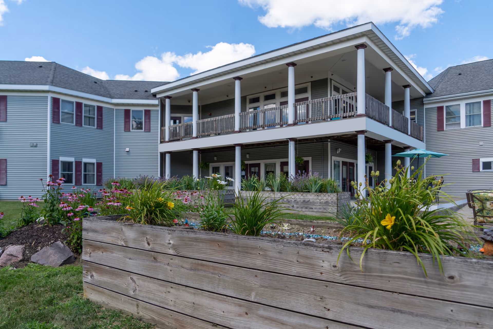 Two-story senior living building with a covered balcony and raised wooden planter boxes filled with flowers.