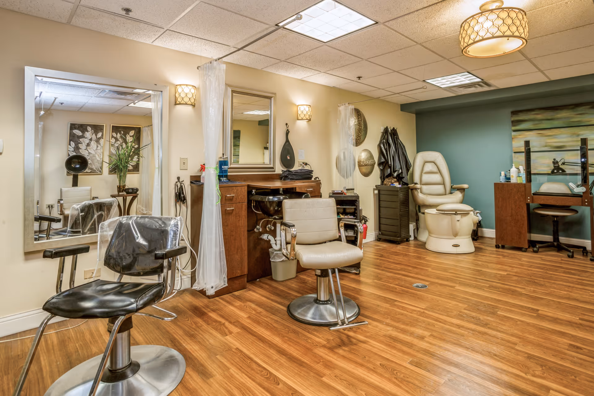 Interior view of a salon area in a senior living facility named Church Creek, featuring salon chairs, a large mirror, a hair washing station, a pedicure chair, and various salon equipment against beige and teal walls with wooden flooring.