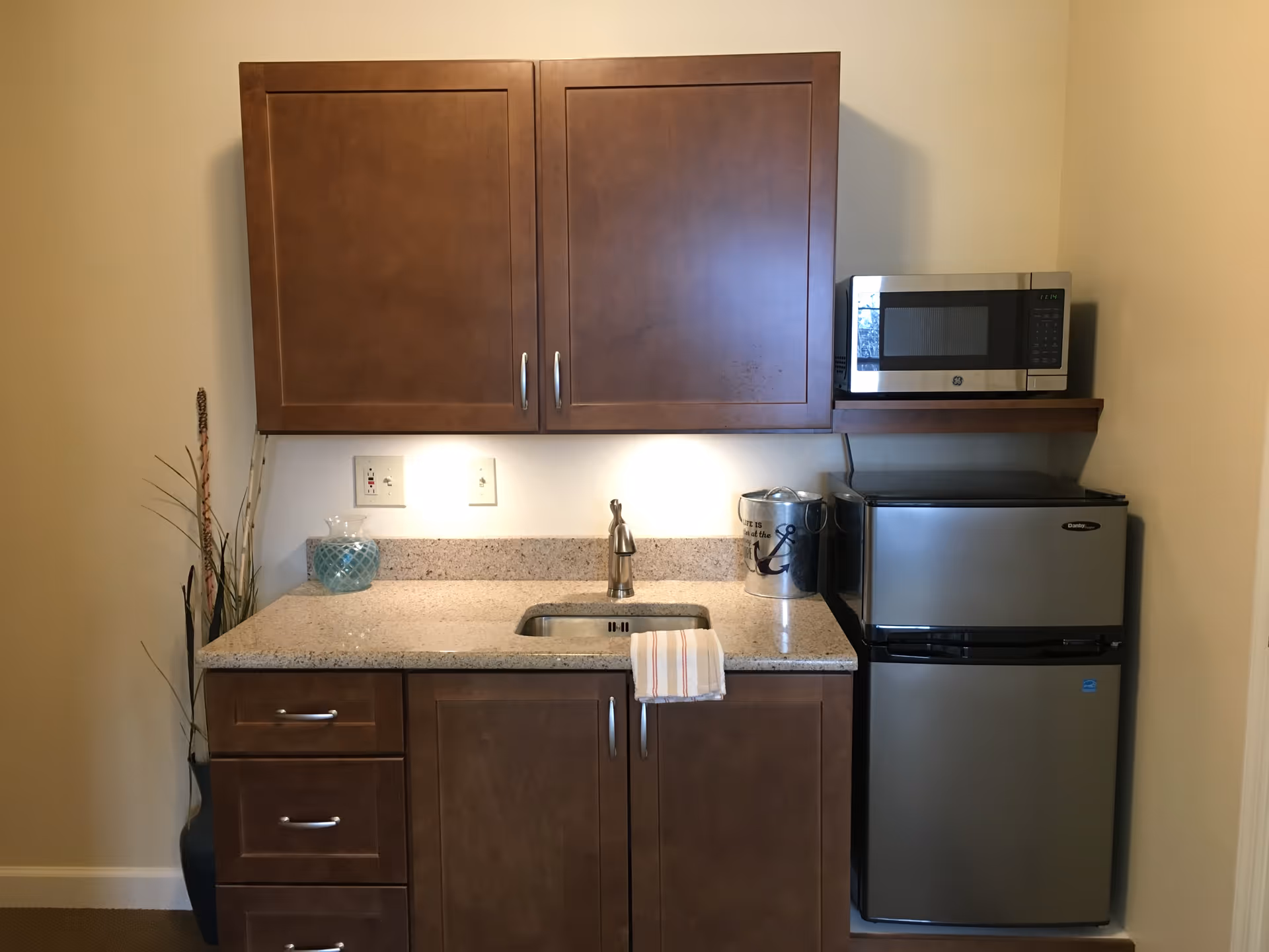 Small kitchenette with wooden cabinets, a sink, a microwave on a shelf, and a stainless mini refrigerator.