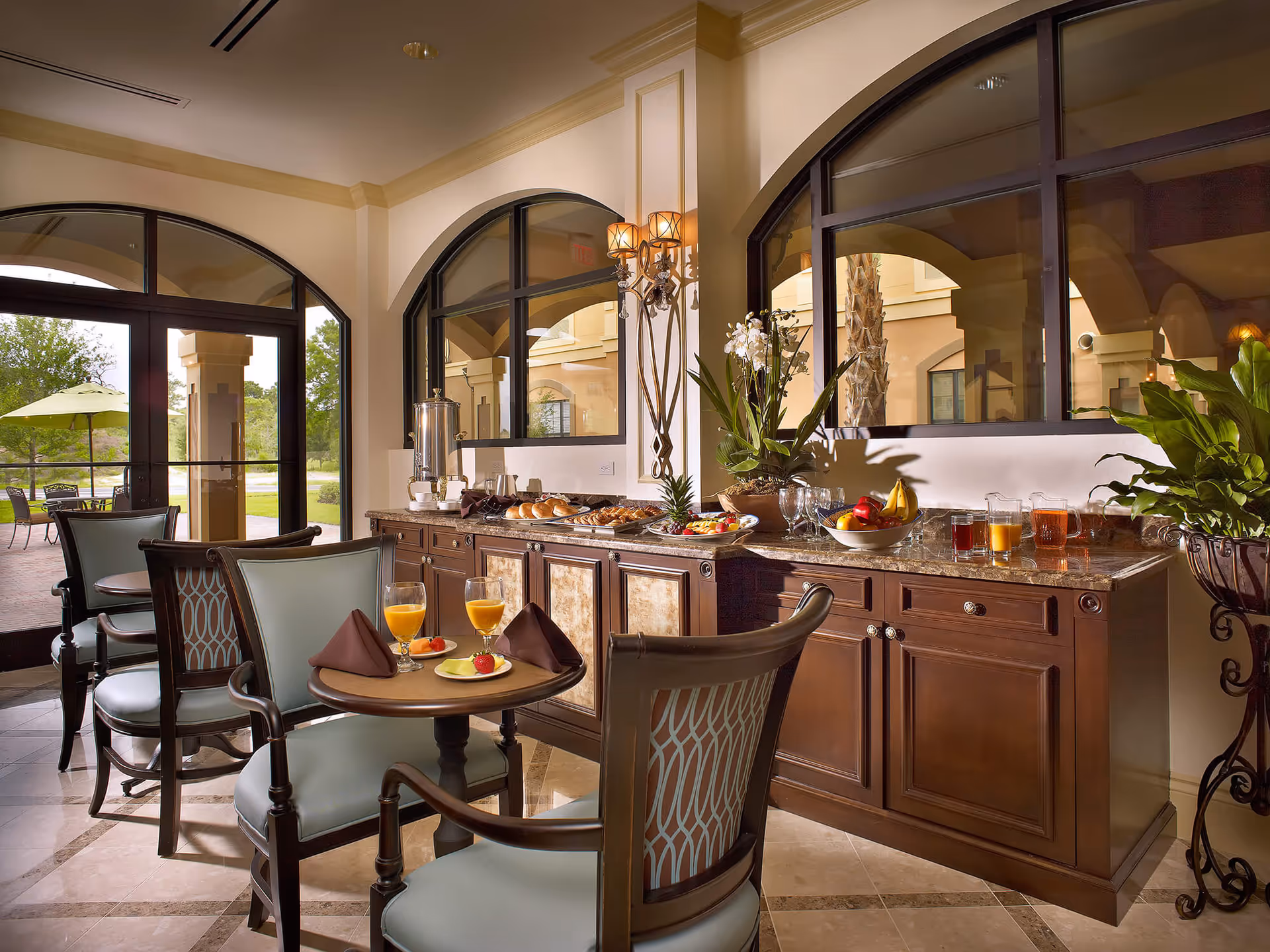 Interior dining area with a buffet counter, small round tables and chairs set with juice and pastries beneath large arched windows.