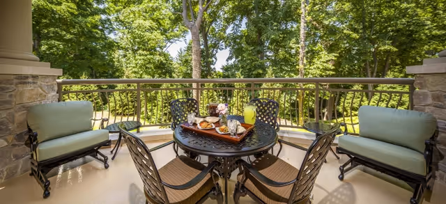 Outdoor patio area with a round black metal table set with a tray of food and drinks, surrounded by four chairs with cushions. Two cushioned benches are positioned on either side of the patio, with a stone pillar on each side. The background shows lush green trees and a sunny sky.