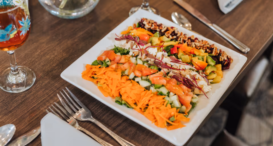 A square white plate with a colorful salad arranged in neat rows including shredded carrots, diced cucumbers and tomatoes, crumbled cheese, chopped bell peppers, and nuts, placed on a wooden table with silverware and a glass of iced tea nearby.