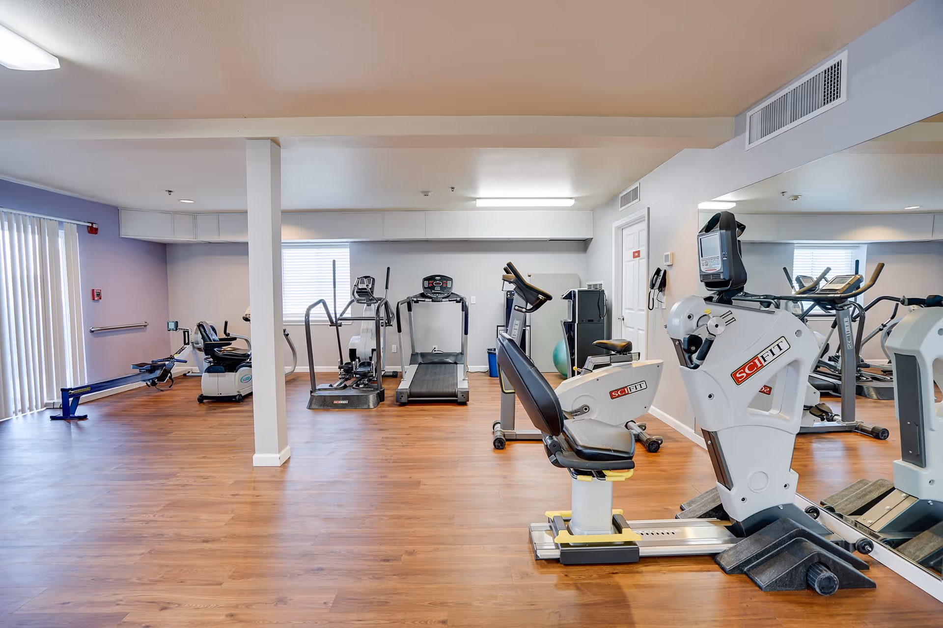 A fitness room with various exercise equipment including a recumbent bike, treadmill, rowing machine, and elliptical machines. The room has wooden flooring, light-colored walls, a large mirror on one side, and a window with blinds letting in natural light.