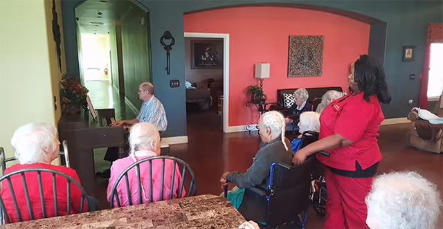 A group of elderly residents in a common room listening to a man playing a piano while a caregiver in red scrubs stands nearby.