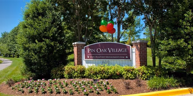 Entrance sign for Pin Oak Village surrounded by landscaped greenery and flowers, with balloons tied to the signpost and a clear blue sky in the background.