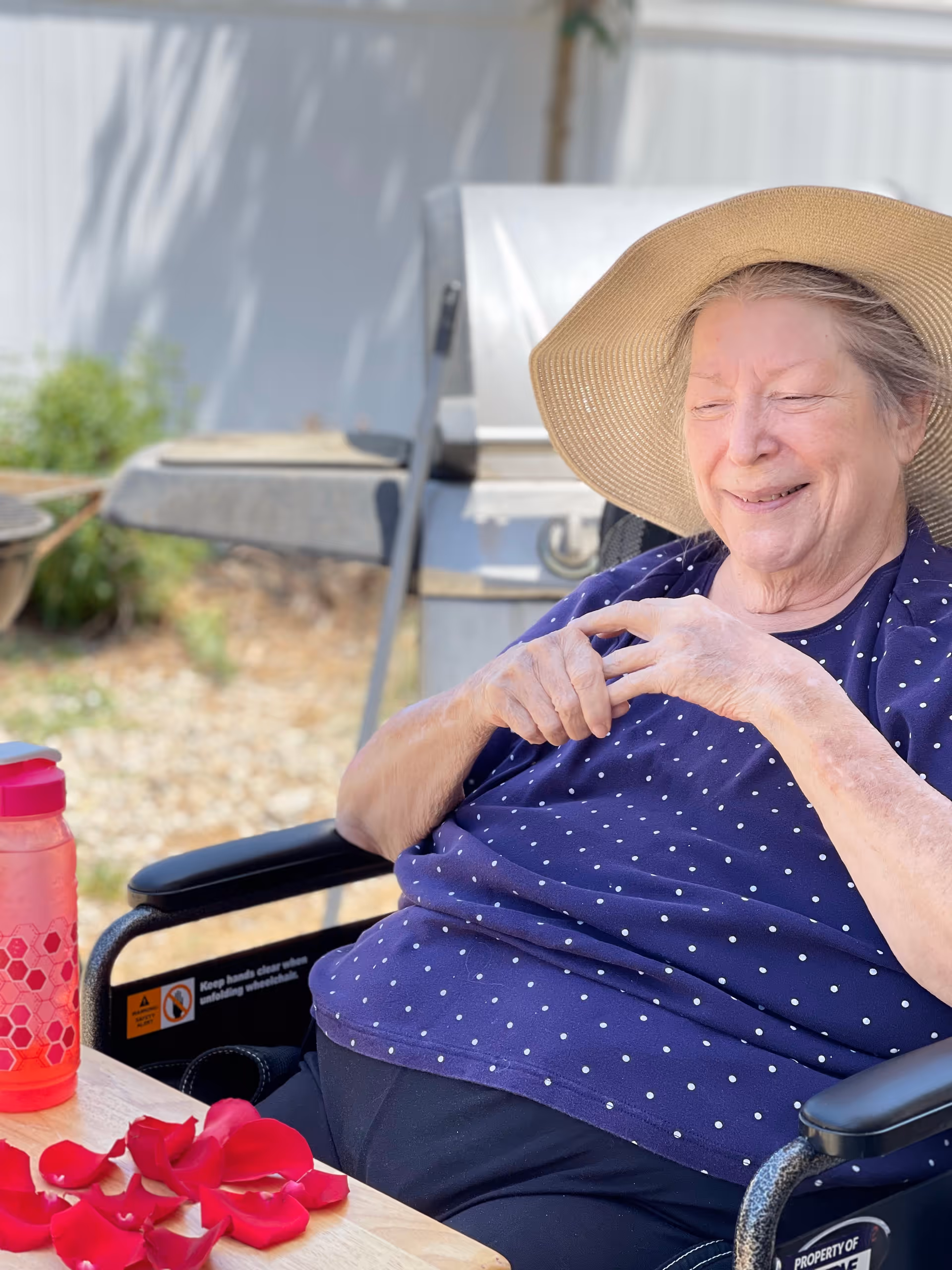 An elderly woman wearing a wide-brimmed hat and a navy blue polka dot shirt sits in a wheelchair outdoors near a table with red rose petals and a pink water bottle. A barbecue grill and garden area are visible in the background.