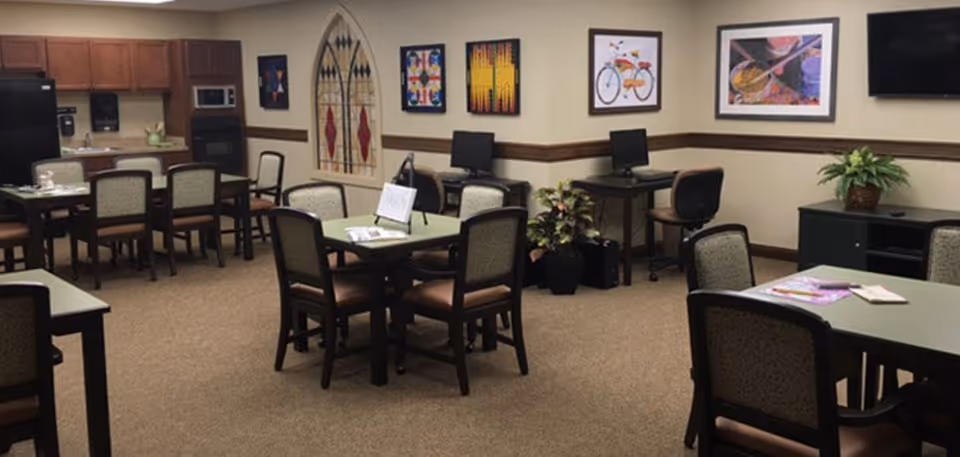 A communal room with several tables and chairs arranged for group seating. The room features a small kitchenette area with a refrigerator, microwave, and sink. There are two computer workstations along one wall, decorative artwork hanging on the walls, and a potted plant near the computers. The carpeted floor and neutral wall colors create a warm and inviting atmosphere.