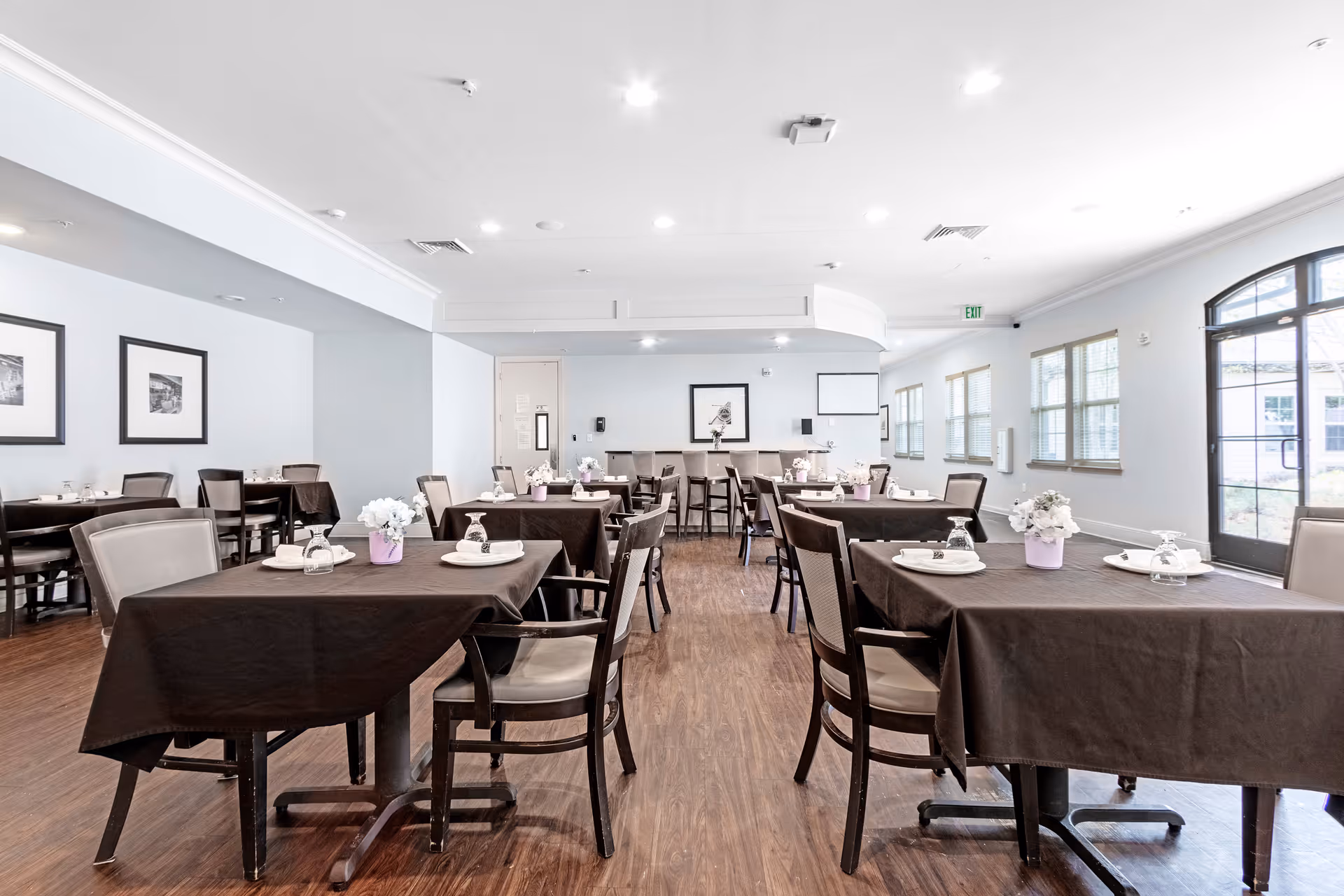 A bright dining room with several tables covered in dark tablecloths, each set with plates, glasses, and small flower arrangements. The room has wooden flooring, light-colored walls, framed pictures, and large windows letting in natural light. There is a door with glass panels on the right side.