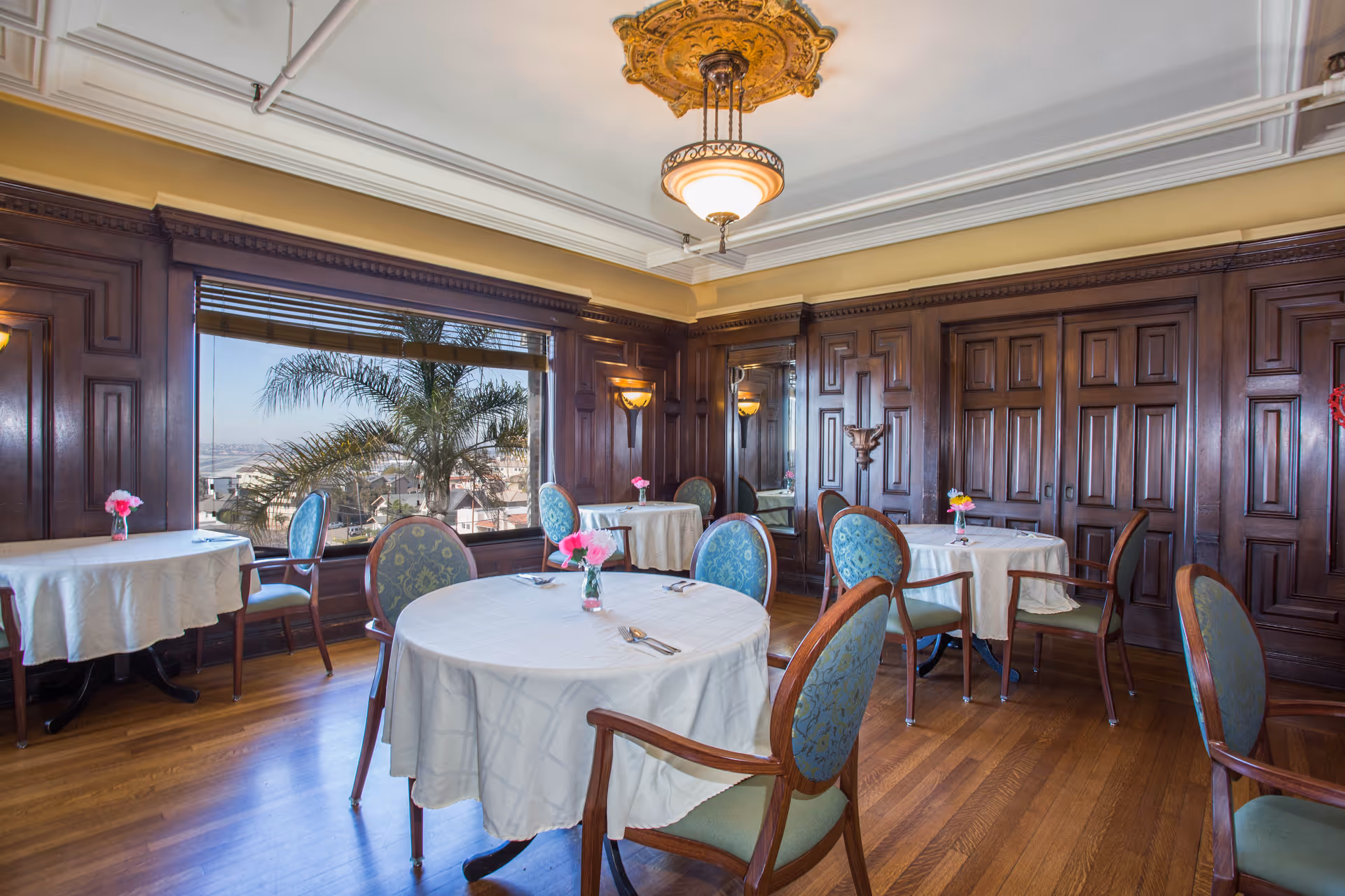 A dining room with round tables covered in white tablecloths, each set with utensils and small vases of pink flowers. The room features wooden paneled walls, large windows with a view of palm trees and a cityscape outside, and an ornate ceiling light fixture.