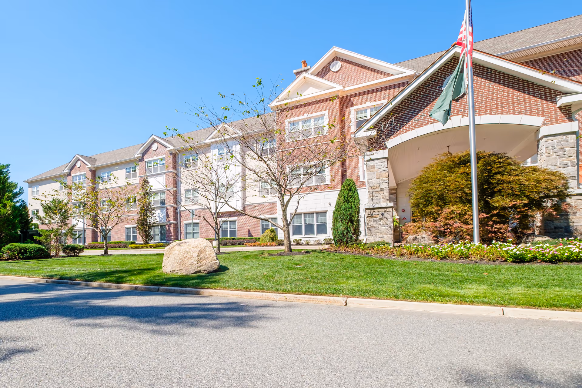 Exterior view of a senior living facility named Brandywine Mahwah by Monarch, showing a large brick building with multiple windows, a well-maintained lawn, trees, shrubs, and a flagpole with an American flag in front under a clear blue sky.