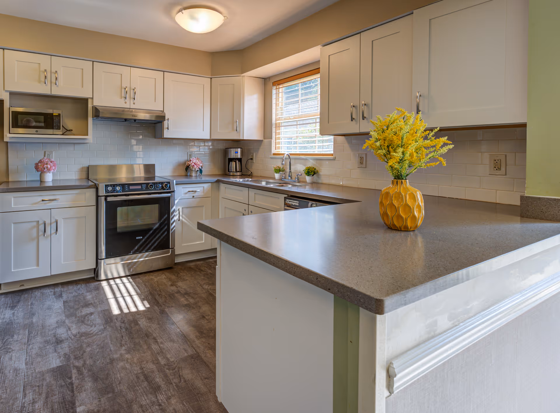 A modern kitchen with white cabinetry, stainless steel appliances including a stove and microwave, a coffee maker, and a large countertop with a yellow vase holding yellow flowers. There is a window above the sink allowing natural light to brighten the space, and the floor has a wood-like finish.