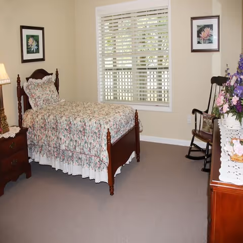 A cozy bedroom with a single wooden bed covered in floral bedding. The room has beige walls, a window with white blinds, two framed floral pictures on the walls, a wooden nightstand with a lamp, a wooden rocking chair, and a dresser with a lace runner and a floral arrangement.