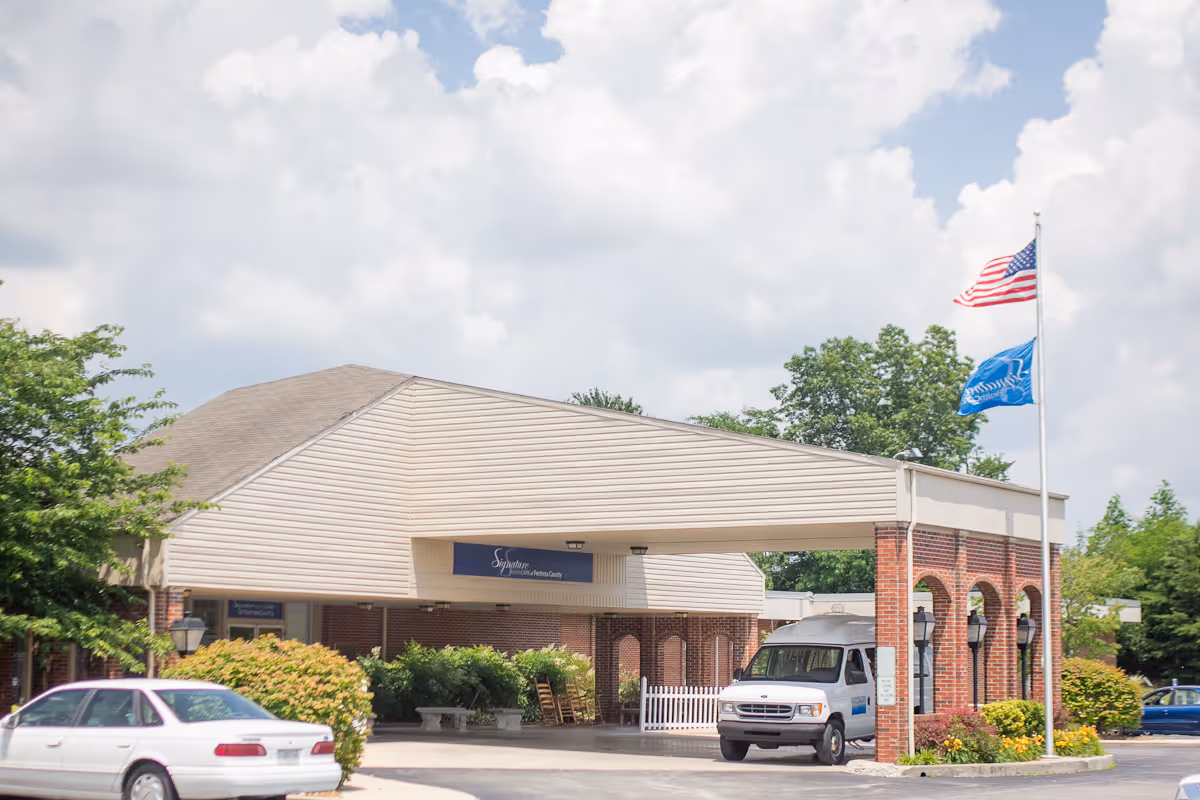 Front entrance of a senior care facility with a covered drop-off, parked vehicles, brick arches, and flagpoles.