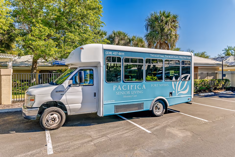 A blue and white senior living shuttle bus parked in a parking lot near a building with palm trees and greenery in the background. The bus has the text 'PACIFICA SENIOR LIVING Memory Care FORT MYERS' and a phone number on the side.