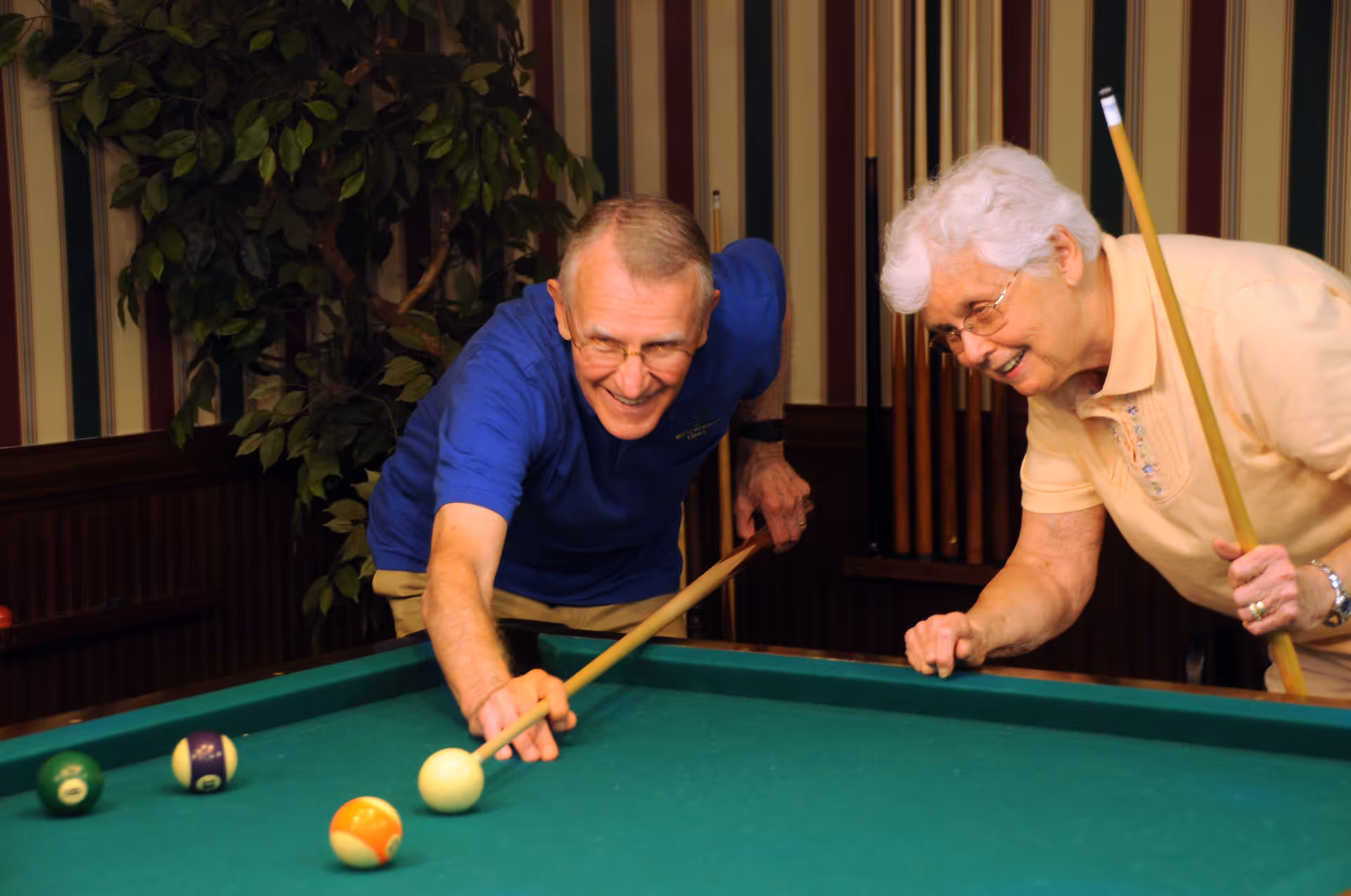 An elderly man and woman playing pool together indoors. The man is aiming a shot with a cue stick while the woman watches and smiles, holding her own cue stick. The room has striped wallpaper and a rack of pool cues on the wall behind them.