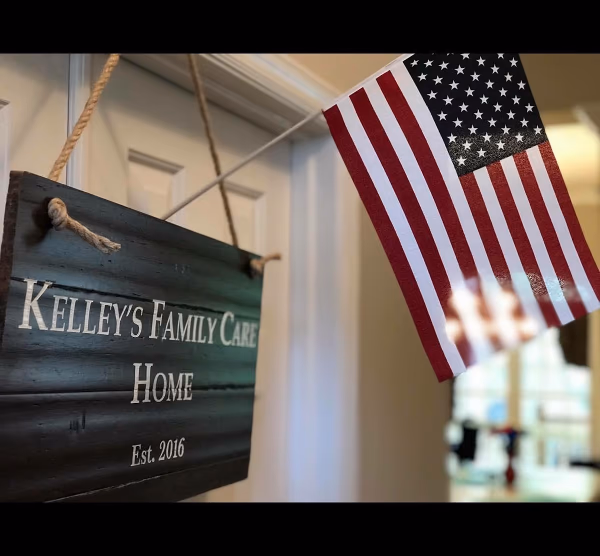 A close-up view of a hanging wooden sign that reads 'Kelley's Family Care Home Est. 2016' next to a small American flag mounted on a pole indoors.