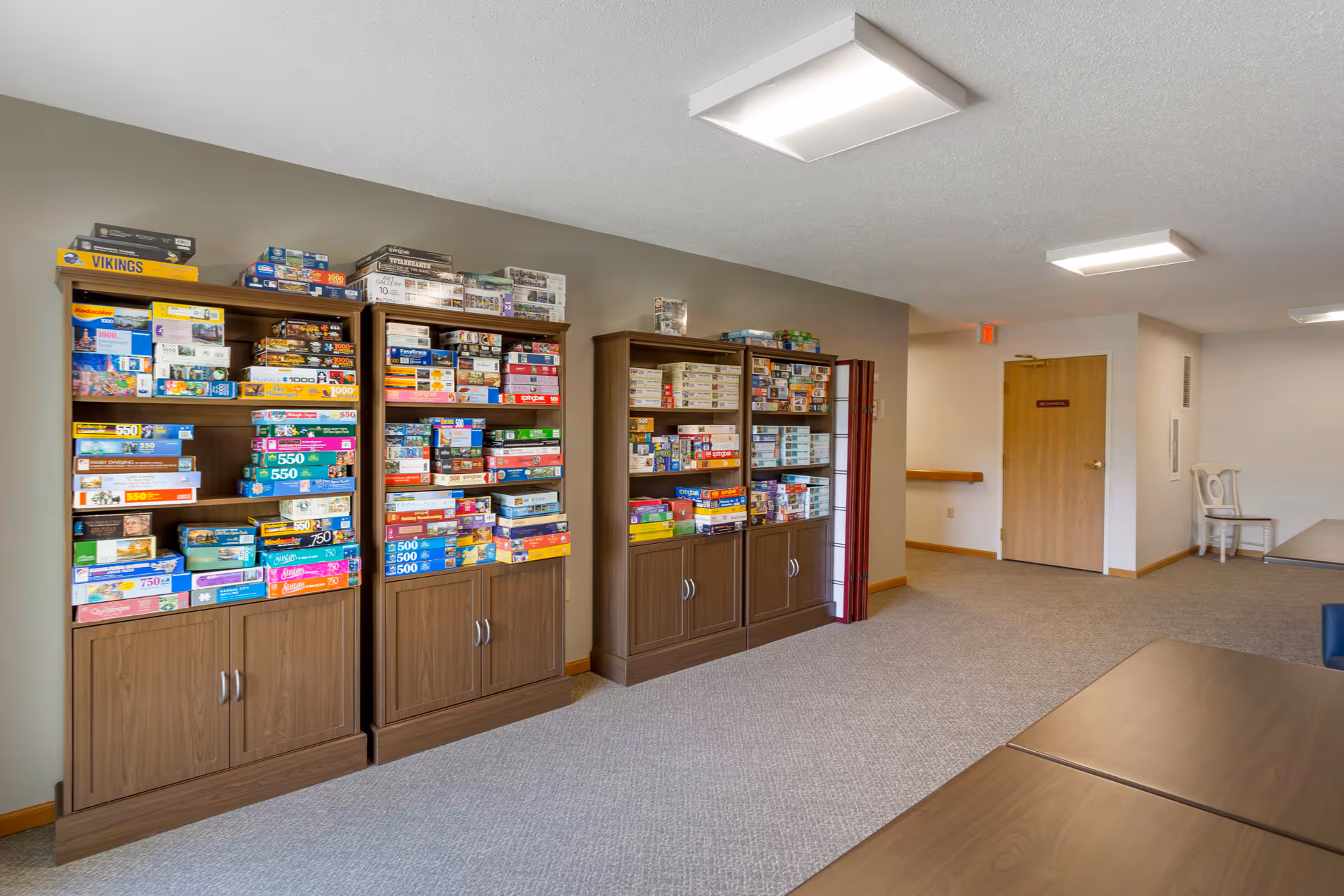 A spacious room with three wooden shelves filled with various board games and puzzles. The room has beige walls, carpeted floor, and ceiling lights. There are tables and chairs visible on the right side, suggesting a recreational or activity area.